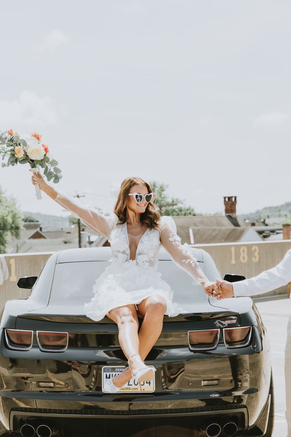 A woman in a white dress poses joyfully on the hood of a black Camaro with a bouquet.