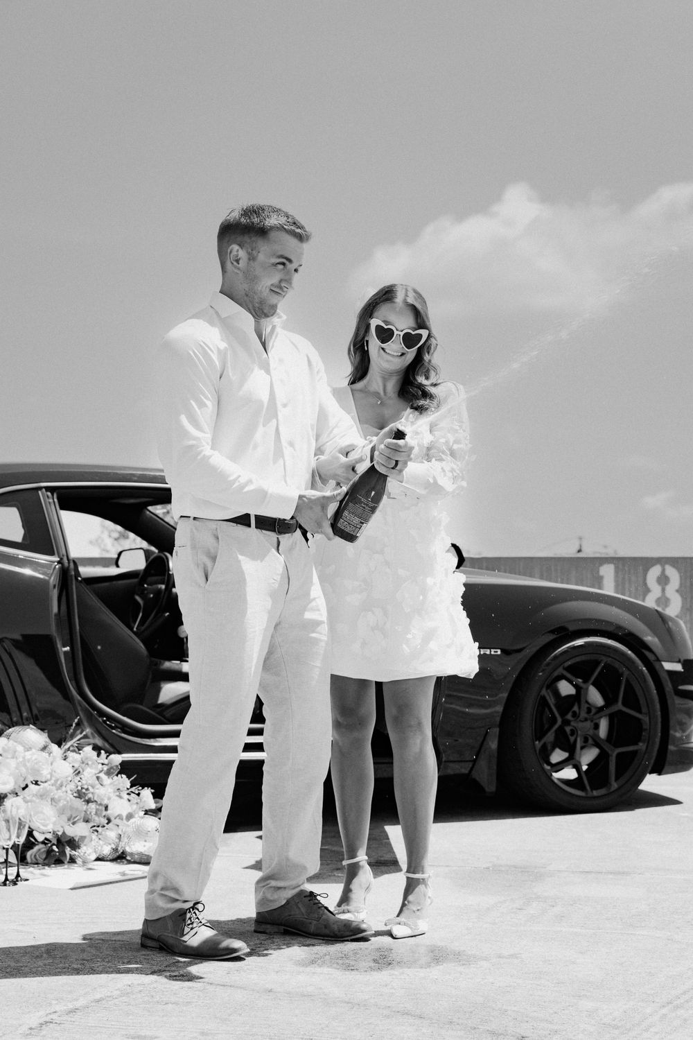 A couple in white outfits stands beside a black car, celebrating with champagne.
