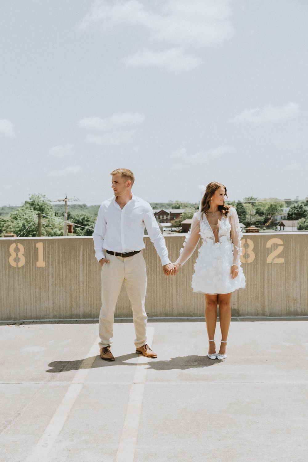 A couple in coordinating white outfits holding hands while standing on a rooftop parking structure.