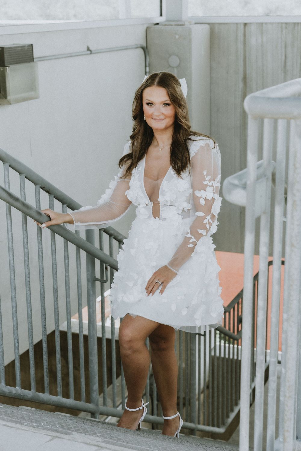 A woman poses on industrial stairs wearing a short white dress with sheer lace sleeves.