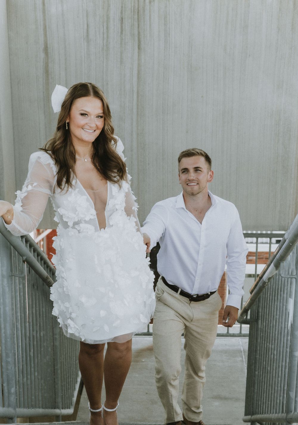 A couple on a stairwell, she in a short white dress with lace details, he in casual white attire.