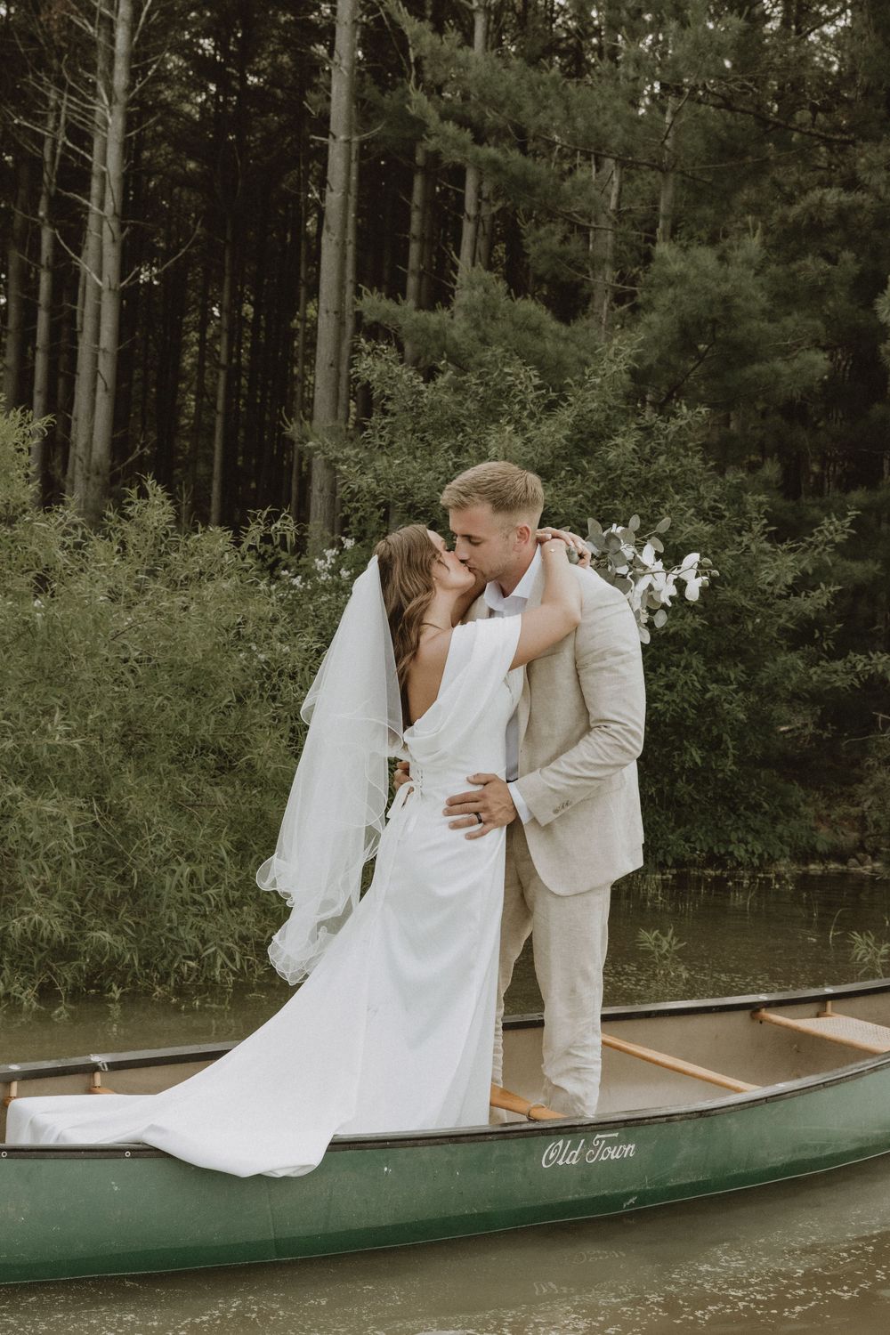 A romantic wedding photoshoot featuring a couple embracing in a green canoe against a wooded forest backdrop.