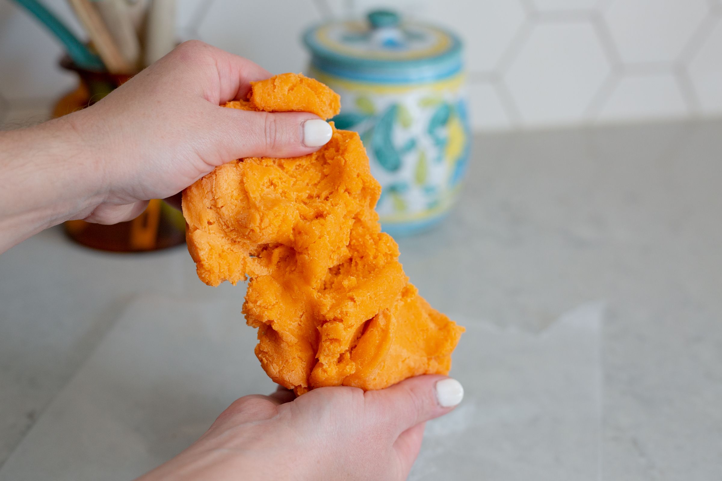 Hands kneading bright orange dough with blue cookie jar in background.