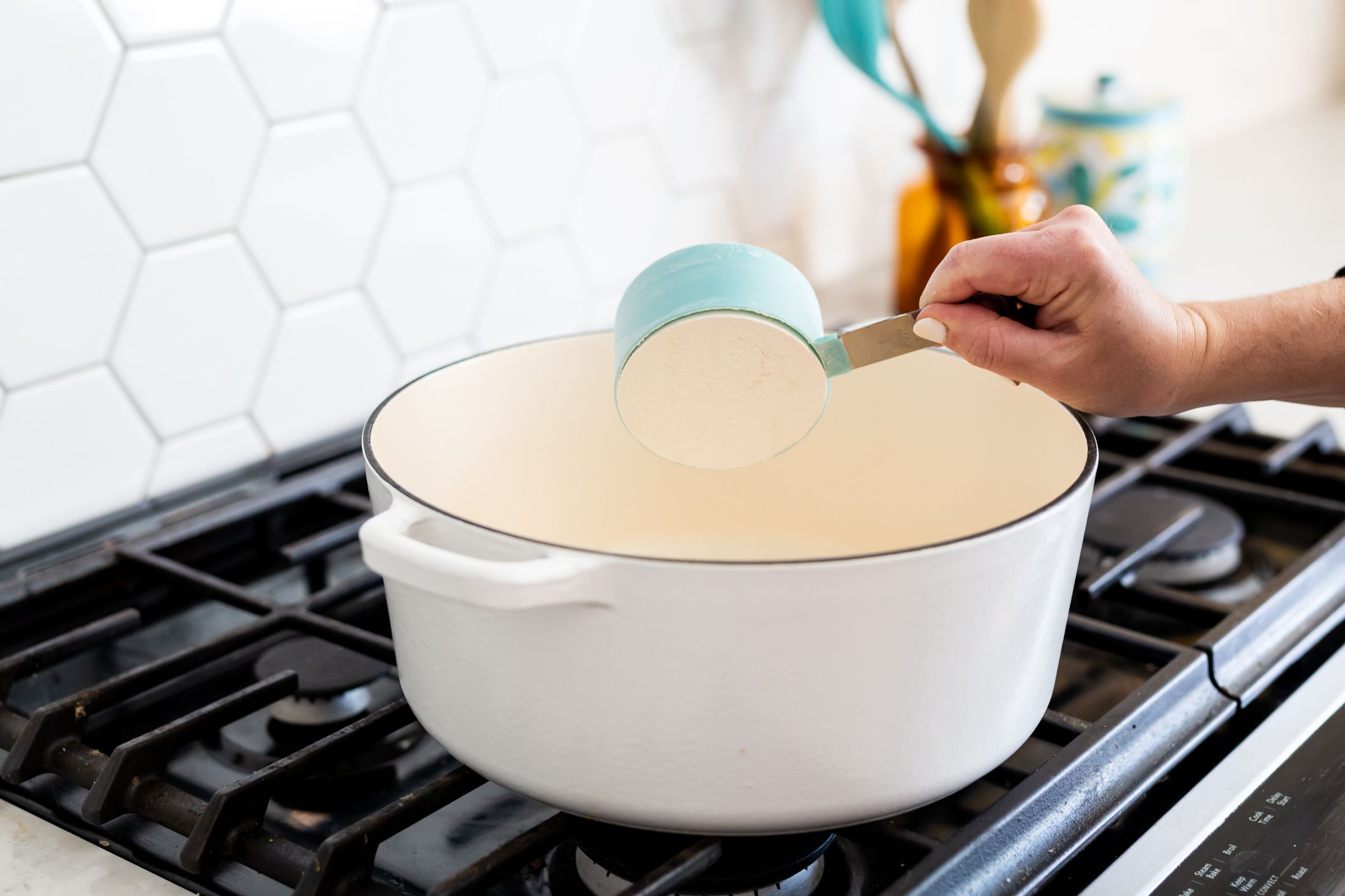 Measuring ingredients into large white Dutch oven on gas stovetop.