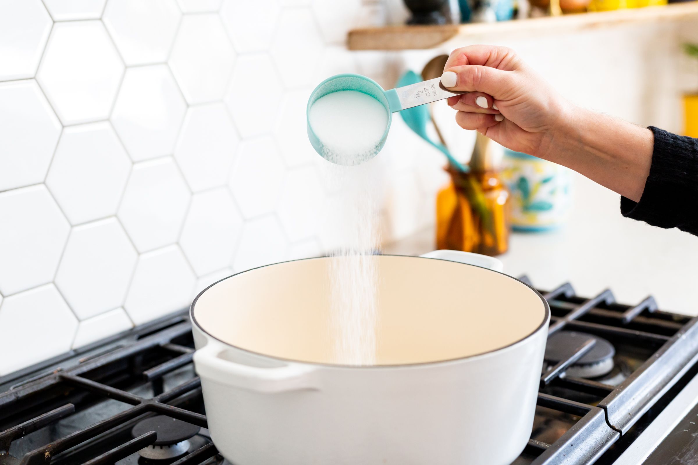 Hand pouring ingredients into white enamel pot on gas stovetop.