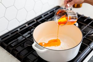 Pouring orange mixture from measuring cup into white pot on stovetop.