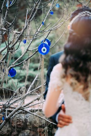 Blue evil eye decorations hang from tree branches with bridal dress in soft focus.