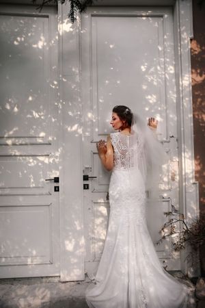 A bride in a lace wedding dress stands before white doors with dappled sunlight creating a romantic atmosphere.