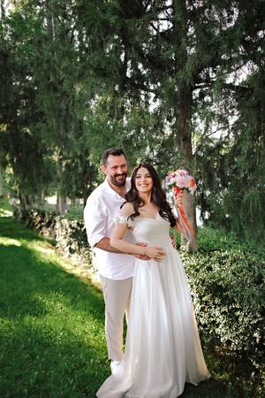 A couple in white attire poses for a wedding photo among evergreen trees.
