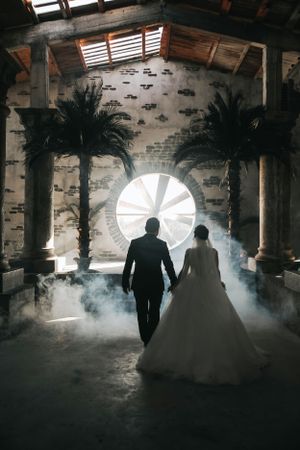 Silhouetted couple walks through palm-lined venue with dramatic backlight and fog effects.