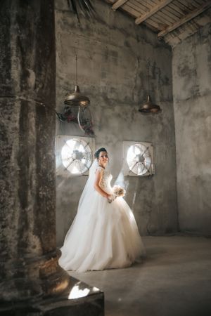 Bride in ballgown poses dramatically in industrial space with circular windows and dramatic lighting.