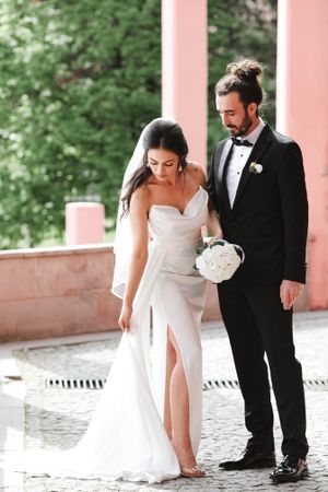 Elegant couple walks together on pink terrace, bride in flowing white gown with bouquet.