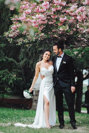 A romantic wedding photo under blooming cherry blossoms with a classic white dress and black tuxedo.