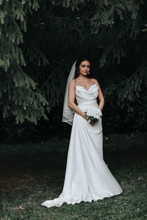 Elegant bride in white satin gown with thigh slit poses against evergreen backdrop.