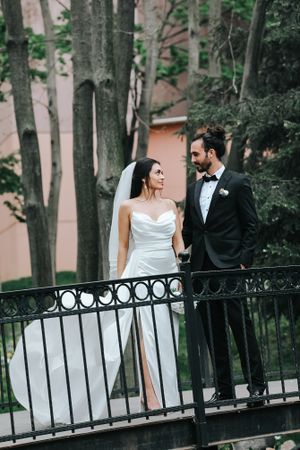 Elegant wedding couple standing together on an ornate black metal bridge surrounded by trees.