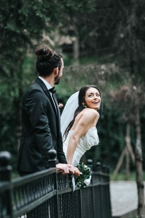 Romantic wedding couple sharing a playful moment on an iron bridge in a forested setting.