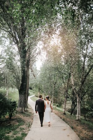 Couple walking hand in hand down a tree-lined path in wedding attire.
