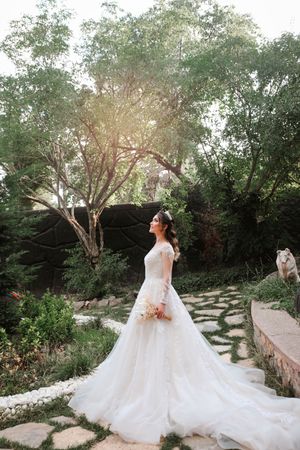 Bride in flowing white gown poses in sunlit garden with trailing dress.