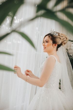 A bride in a sparkly white dress and tiara smiling beside tropical leaves.