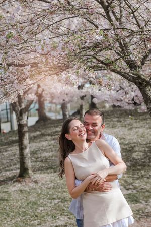 A romantic couple embraces under blooming cherry blossom trees in a springtime park setting.