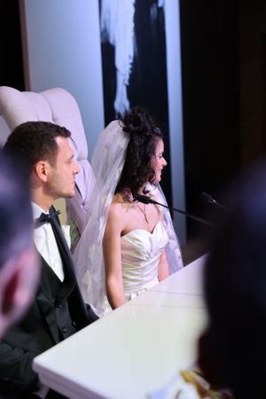 A bride and groom sit together at their wedding reception, with the bride wearing a white strapless gown and veil.