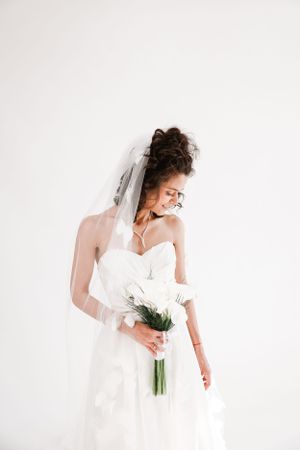 A bride in a white strapless gown and veil holds a simple bouquet while looking down against a white backdrop.