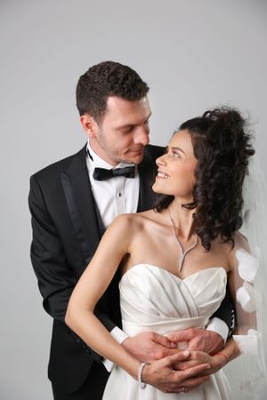 A romantic couple in formal wedding attire embraces while gazing at each other against a light gray background.