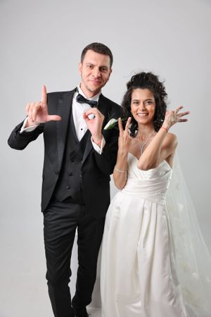 Wedding couple in formal attire making rock and roll hand gestures against gray background.