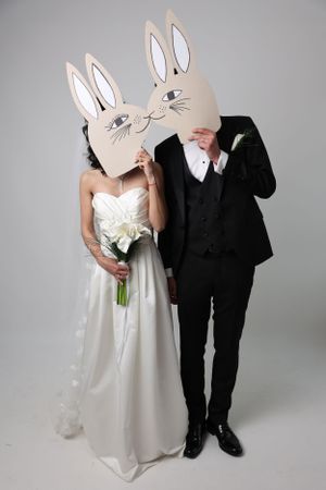 A bride and groom wearing matching white paper bunny masks pose together, with her in a white dress and him in a black suit.