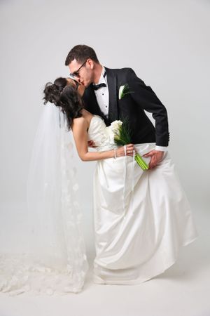 A romantic wedding photo of a couple kissing while the bride holds a white bouquet against a light background.