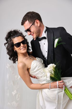 A joyful bride and groom wearing sunglasses share a playful moment while posing with a white bouquet against a light backdrop.