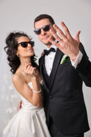 Stylish couple in wedding attire wearing sunglasses showing off rings in a playful pose against gray background.