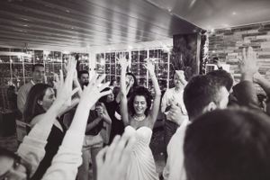A large group of wedding reception guests dance with raised arms on a crowded dance floor in black and white.