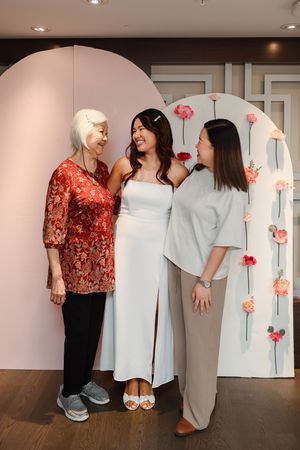 Three people pose together in coordinated white and gray outfits against a pink wall with red floral decorations.