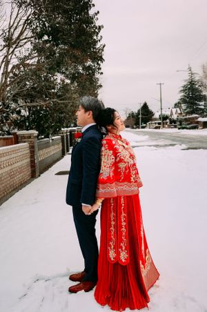 Couple in traditional wedding attire standing back to back on snowy street, bride wearing ornate red Chinese dress.