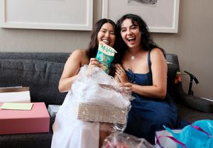Two friends sit on a grey couch sharing gifts and laughing together during an unwrapping session.