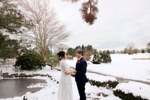 A couple shares a romantic winter wedding moment beside a snow-covered pond with bare trees in the landscape.