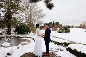 Couple embraces during winter wedding ceremony beside a snowy pond with evergreen trees in the background.
