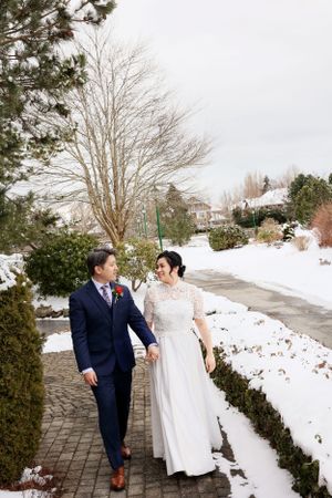 Two people in wedding attire walk together on a snowy brick path surrounded by winter landscaping.