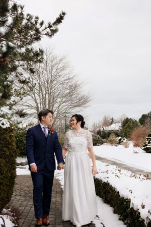 Couple in wedding attire walking along a snow-covered garden path surrounded by winter landscaping.