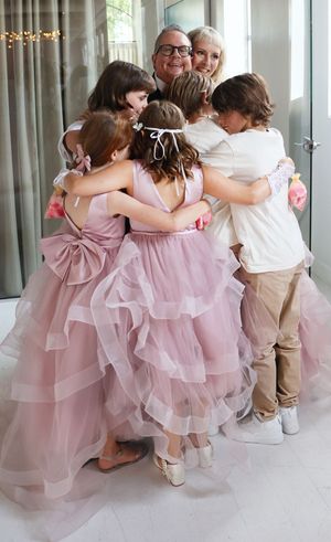Children in pink tulle dresses and khaki outfits sit together in a row against a bright white wall.