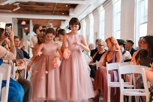 Wedding party in pink bridesmaid dresses gather in a bright white room with exposed wooden beams and large windows.