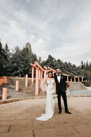 Wedding couple poses in front of pink columns and evergreen trees under cloudy sky.