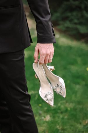 Person in formal wear holds elegant crystal-embellished beige wedding heels against green outdoor background.