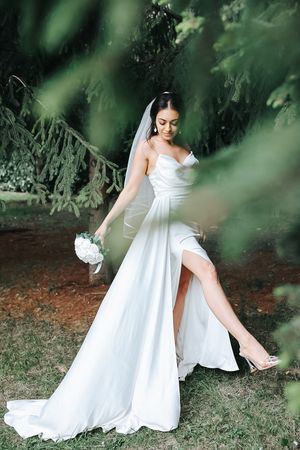 Bride in flowing white gown with high slit poses gracefully in garden setting.