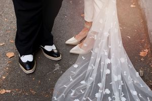 A close-up of wedding shoes showing the groom's black and white dress shoes next to a bride's flowing veil with heart details.