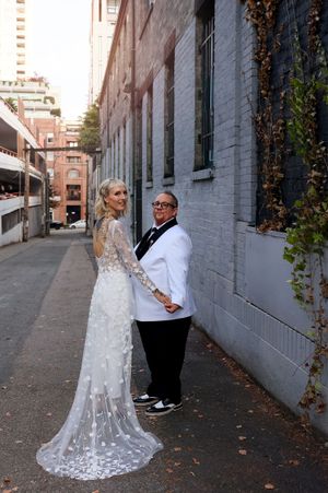 A couple in wedding attire poses in an urban alleyway with brick walls and fallen autumn leaves.