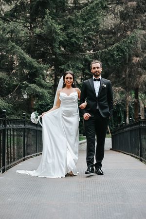 Elegant couple in wedding attire walking on bridge surrounded by evergreen trees.