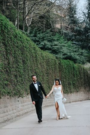 Wedding couple walking hand in hand along path beside tall mossy wall.