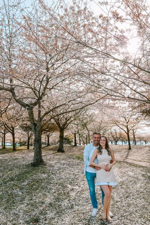 A couple poses beneath blooming cherry blossom trees in a scenic spring park setting.